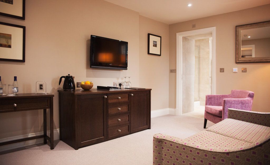 Habitación de hotel con un mueble de madera oscura, un televisor montado en la pared, un sillón rosa y un otoman estampado.