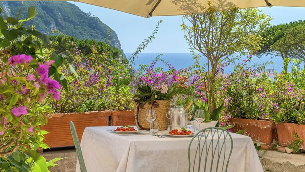 An outdoor dining area with a table set for two, surrounded by lush greenery and vibrant flowers, overlooking a scenic coastal view. - Hotel Villa Melodie - Adults Only (Forio)