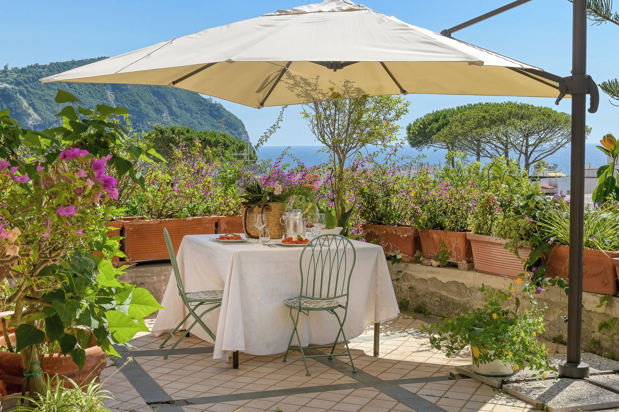 Un área de comedor al aire libre con una mesa dispuesta para dos personas, rodeada de macetas con plantas y vista al mar.
