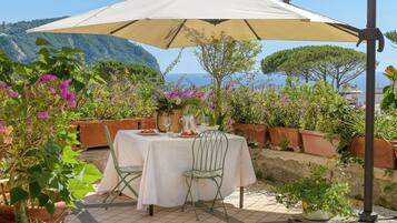 Un área de comedor al aire libre con una mesa dispuesta para dos personas, rodeada de macetas con plantas y vista al mar.