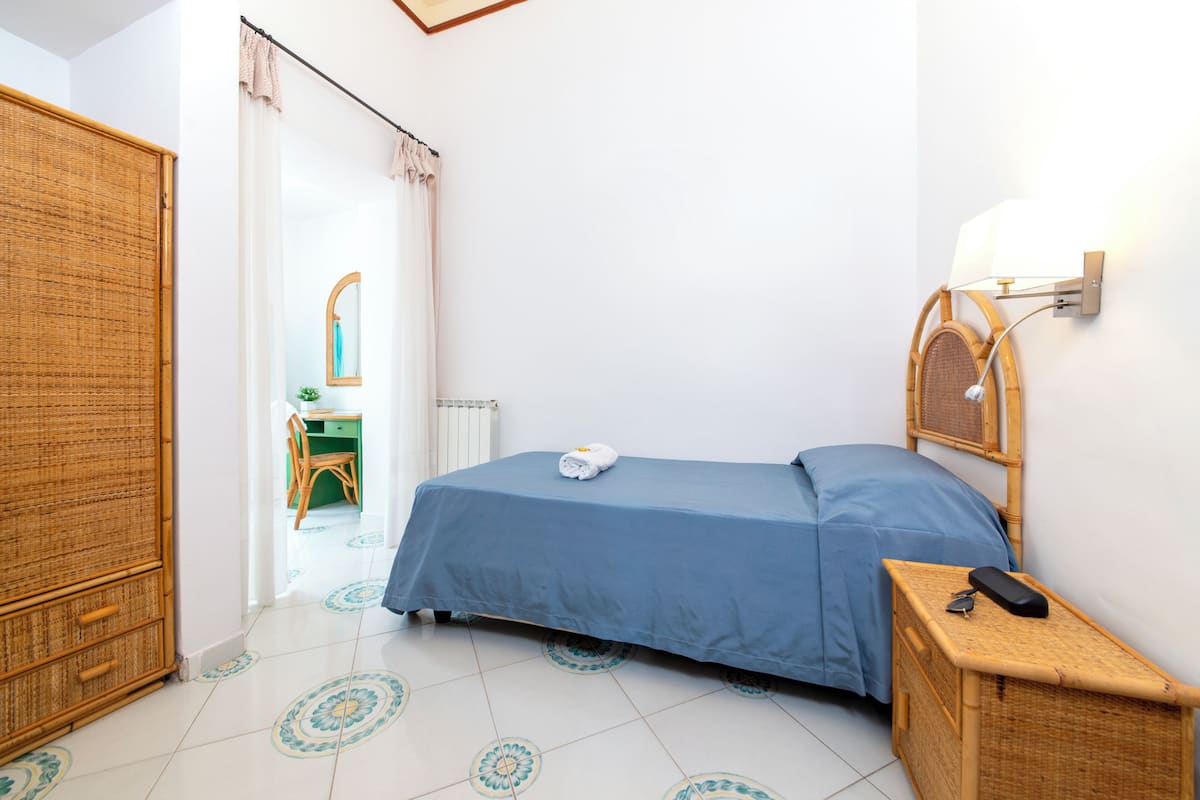 Un dormitorio con cama, una mesita de noche con lámpara, una mesita auxiliar de mimbre y vistas a un baño con espejo y silla.