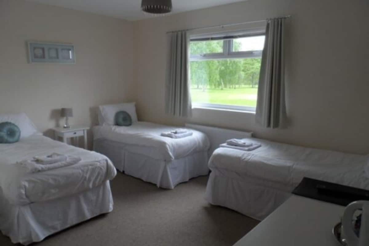 Habitación de hotel con dos camas individuales, una mesita de noche pequeña con lámpara, una ventana con cortinas y vistas a zonas verdes.