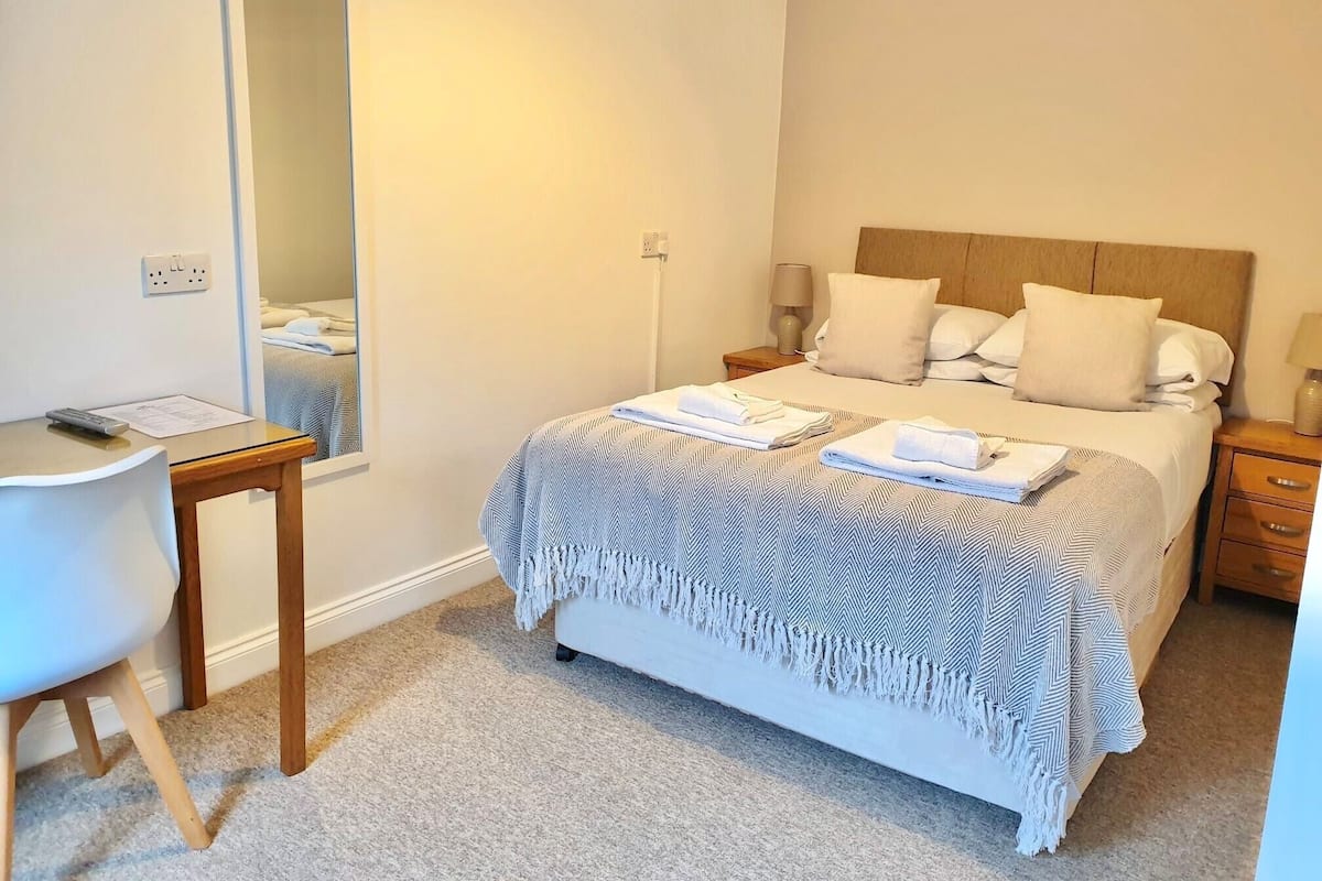 Un dormitorio ordenado con cama, mesitas de noche, escritorio y espejo.