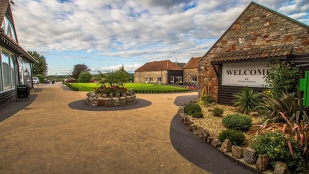 A welcoming entrance with a 'WELCOME' sign, a paved driveway, and a landscaped garden area. - Mendip Spring Golf Club (Bristol)