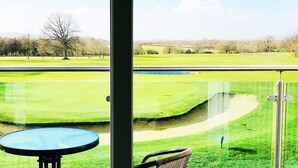 A view from a balcony with a glass railing, overlooking a well-maintained golf course with a pond and trees in the background. - Mendip Spring Golf Club (Bristol)