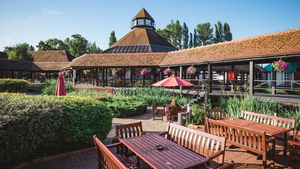 An outdoor patio with wooden tables and benches, surrounded by greenery and a building with a thatched roof. - CIM Business Centre (Maidenhead)