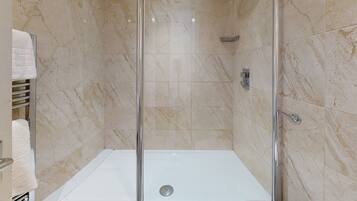 Una ducha sin barreras con puerta de vidrio, azulejos de mármol color crema y suelo blanco.