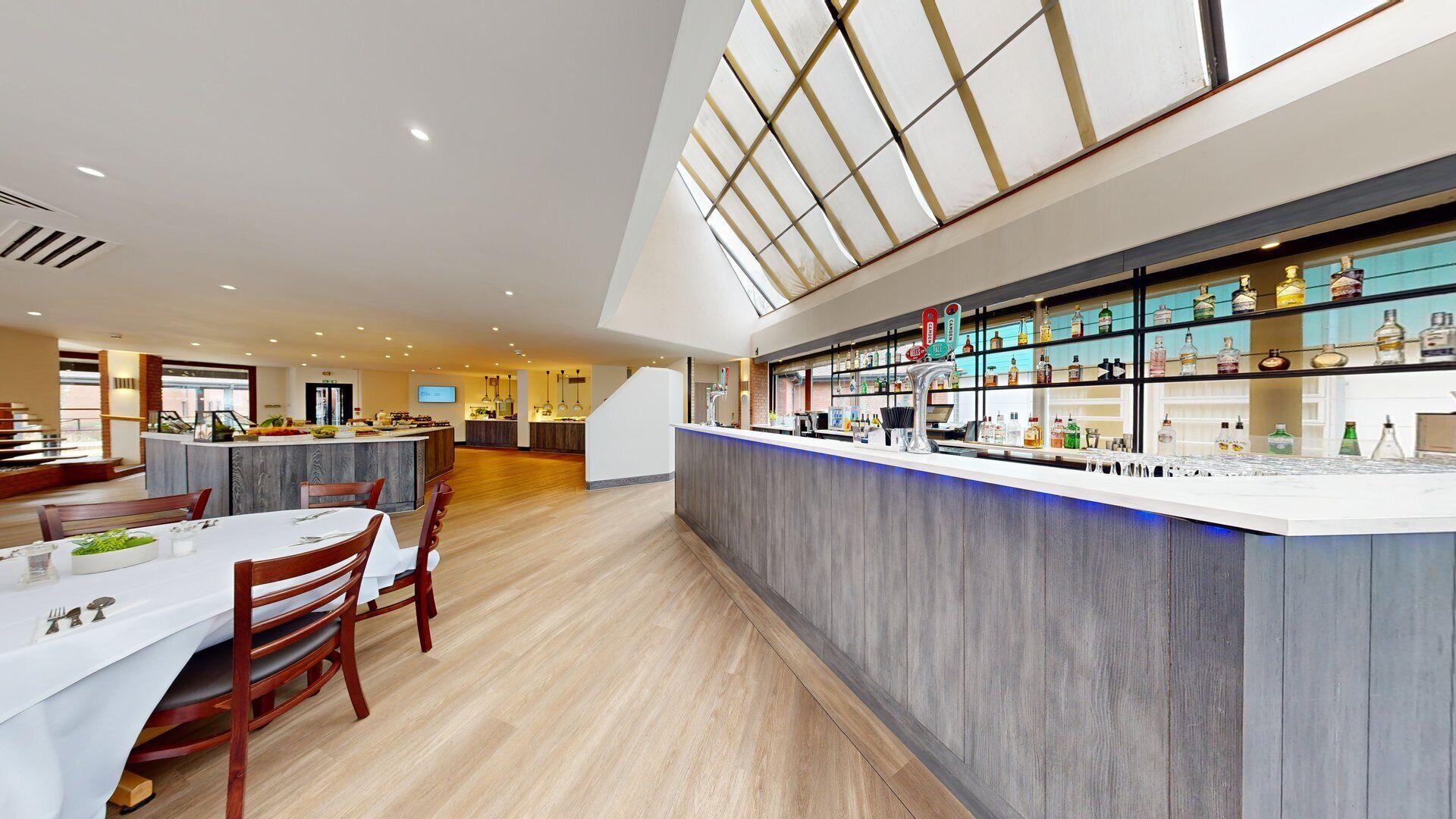 Un restaurante moderno con zona de bar, suelo de madera y un techo alto con tragaluces.