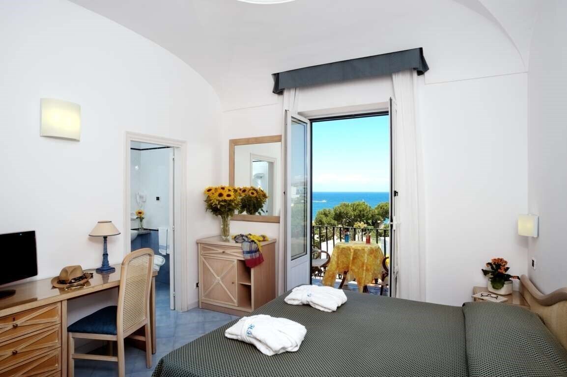 A hotel room with a bed, a desk, a chair, a TV, a window with a view of the sea, and a balcony with a table and chairs.