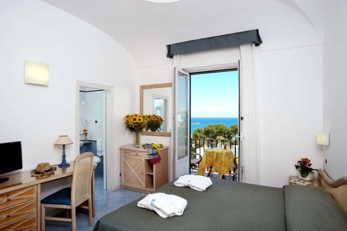 A hotel room with a bed, a desk, a chair, a TV, a window with a view of the sea, and a balcony with a table and chairs.