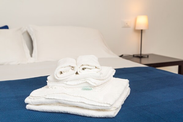 Una cama bien hecha con sábanas blancas y una colcha azul, una mesita de noche con una lámpara y un enchufe de pared.