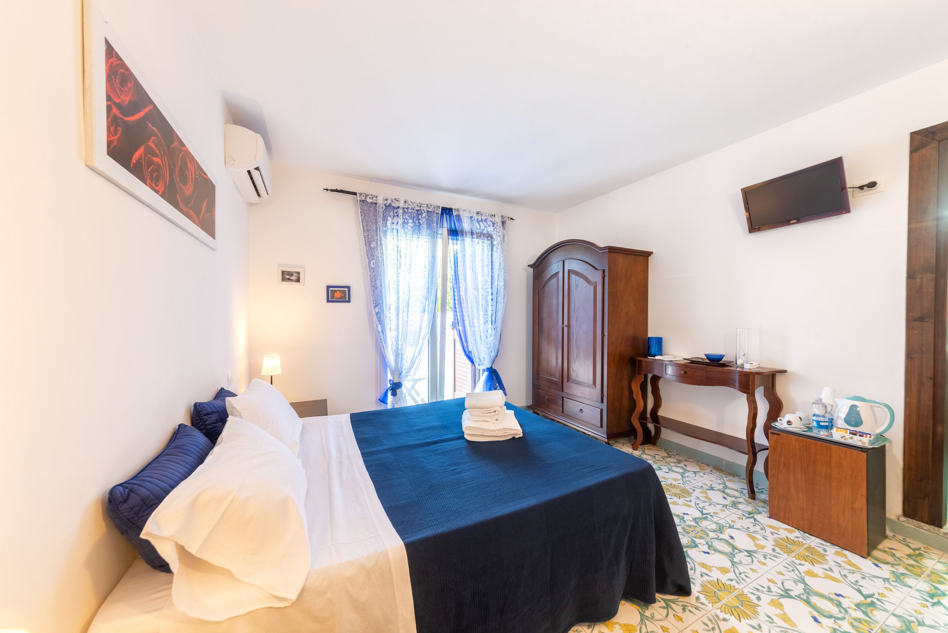 A neatly made bed with a blue bedspread, a wooden wardrobe, a small table with a lamp, and a television on the wall.