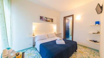 A hotel room with a bed, bedside lamps, a painting, a door, and a shelf with items.