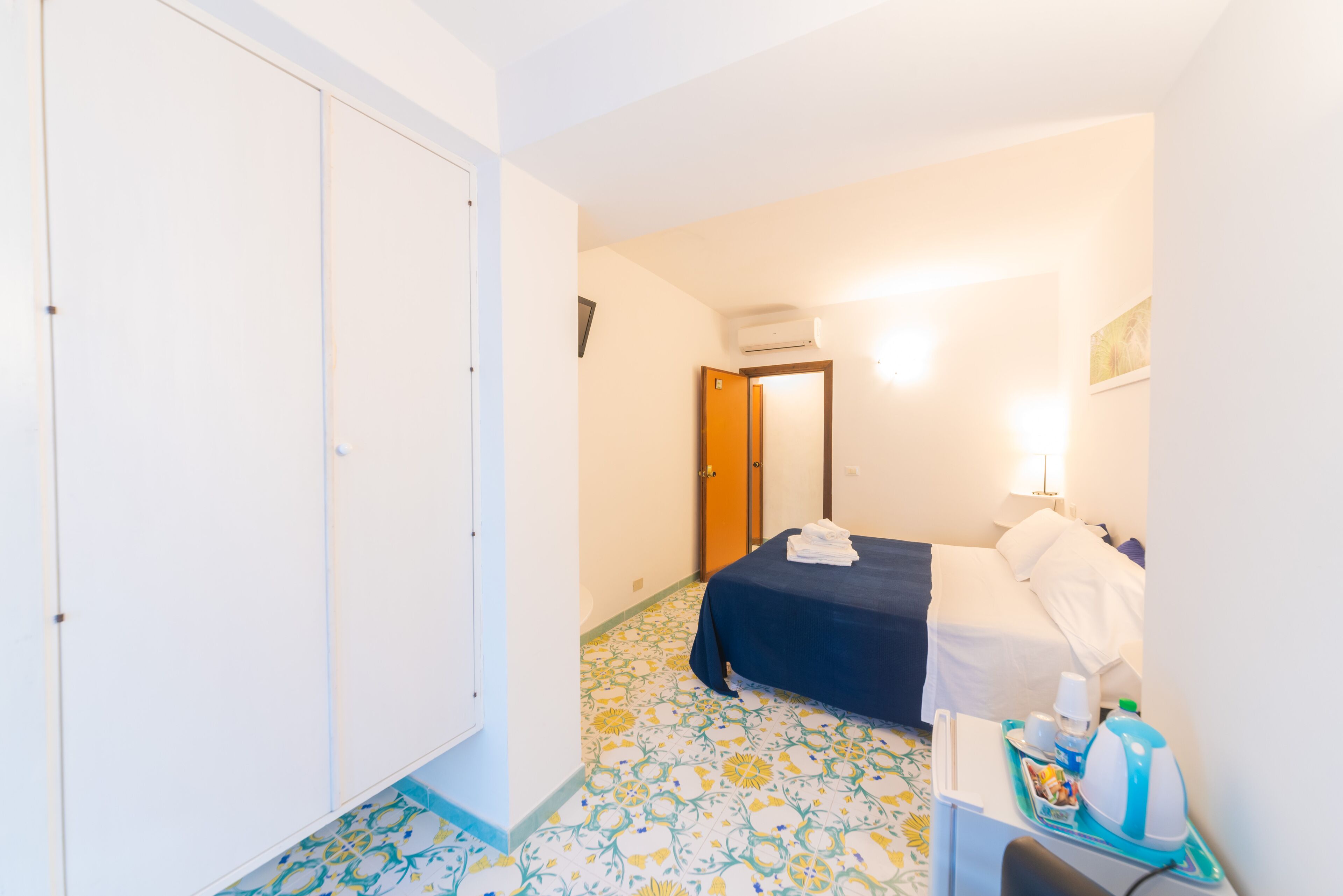 A hotel room with a bed, bedside table, lamp, and a tiled floor.