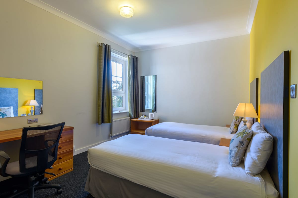 A hotel room with two beds, a desk with a chair, a window with curtains, and a lamp on the nightstand.