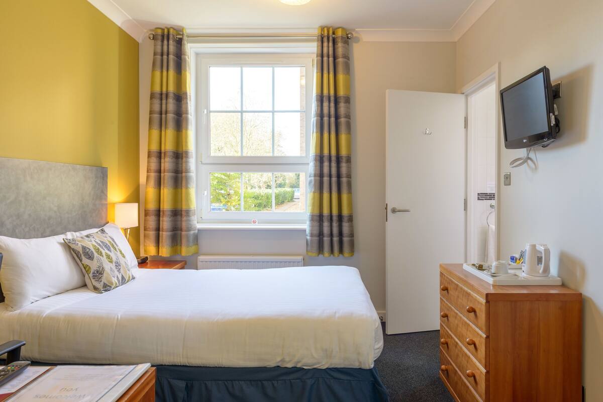 A hotel room with a bed, a wooden dresser, a television, and a window with curtains.