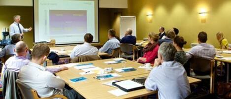 A conference room with a presentation screen, attendees seated at tables, and a whiteboard in the background.