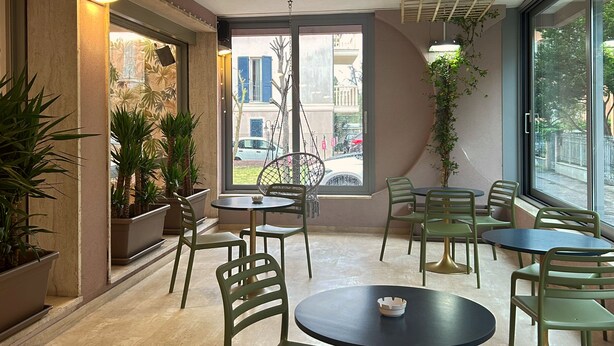 A modern indoor seating area with round tables and green chairs, large windows offering a view of the street, and potted plants adding greenery. - Hotel Concorde (Bellaria-Igea Marina)