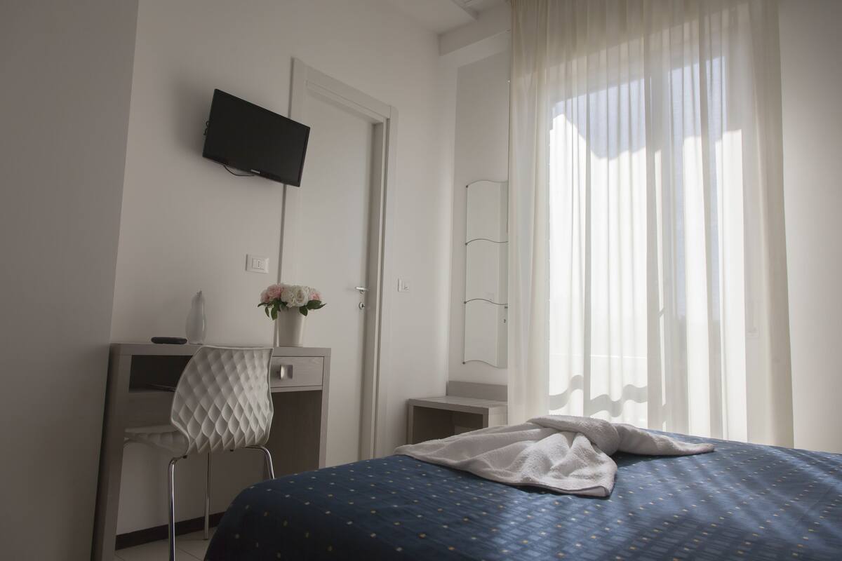 Une chambre à coucher avec un lit, un bureau, une chaise, un téléviseur et une fenêtre avec des rideaux.