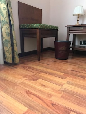 Een hoek van een kamer met een houten vloer, een groene fauteuil, een prullenbak en een lamp op een tafel.