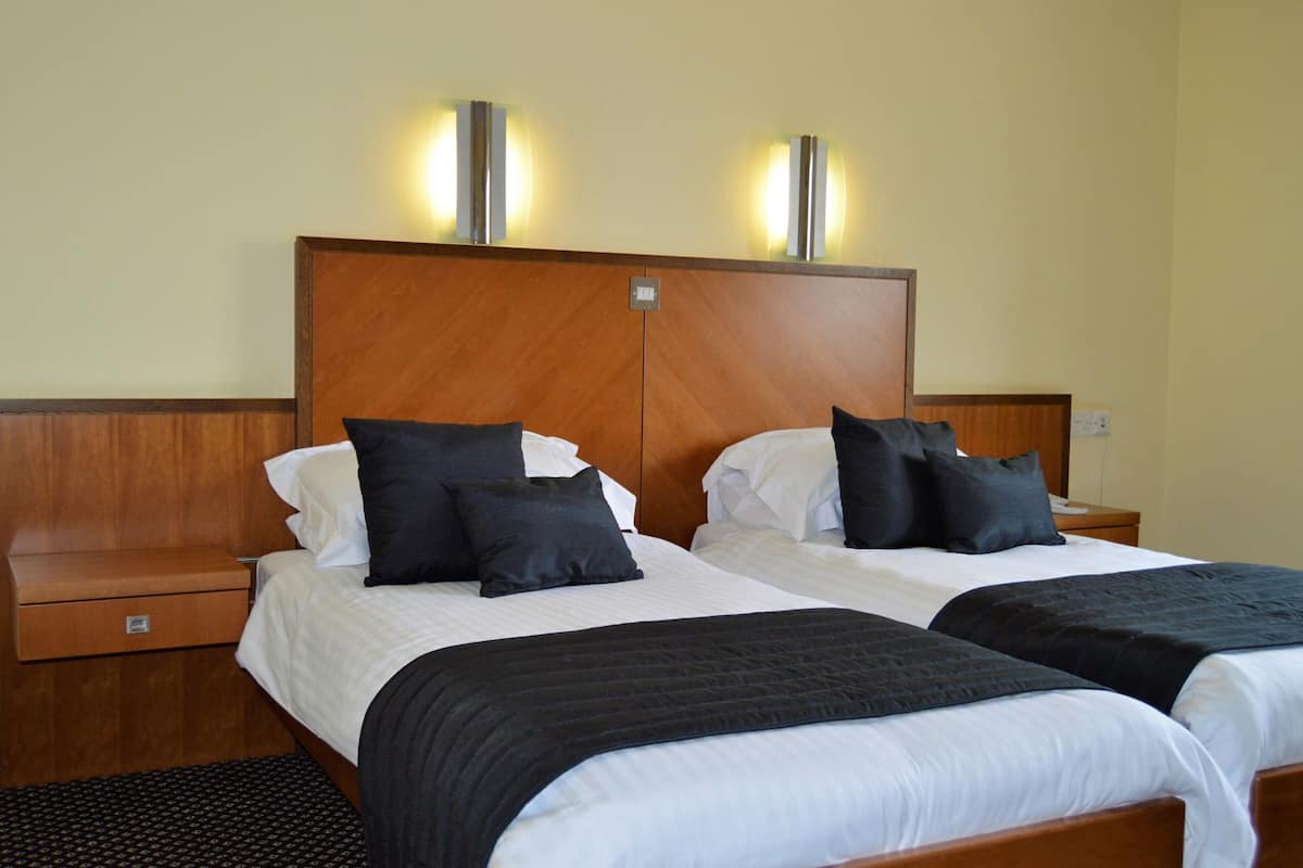 A hotel room with two beds, wooden headboards, bedside tables, and wall-mounted lamps.