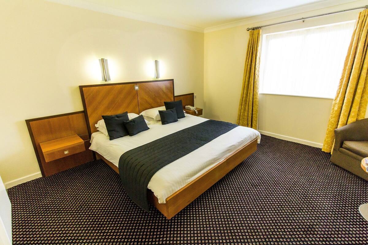 A hotel room with a large bed, a bedside table, a nightstand, a chair, and a window with curtains.