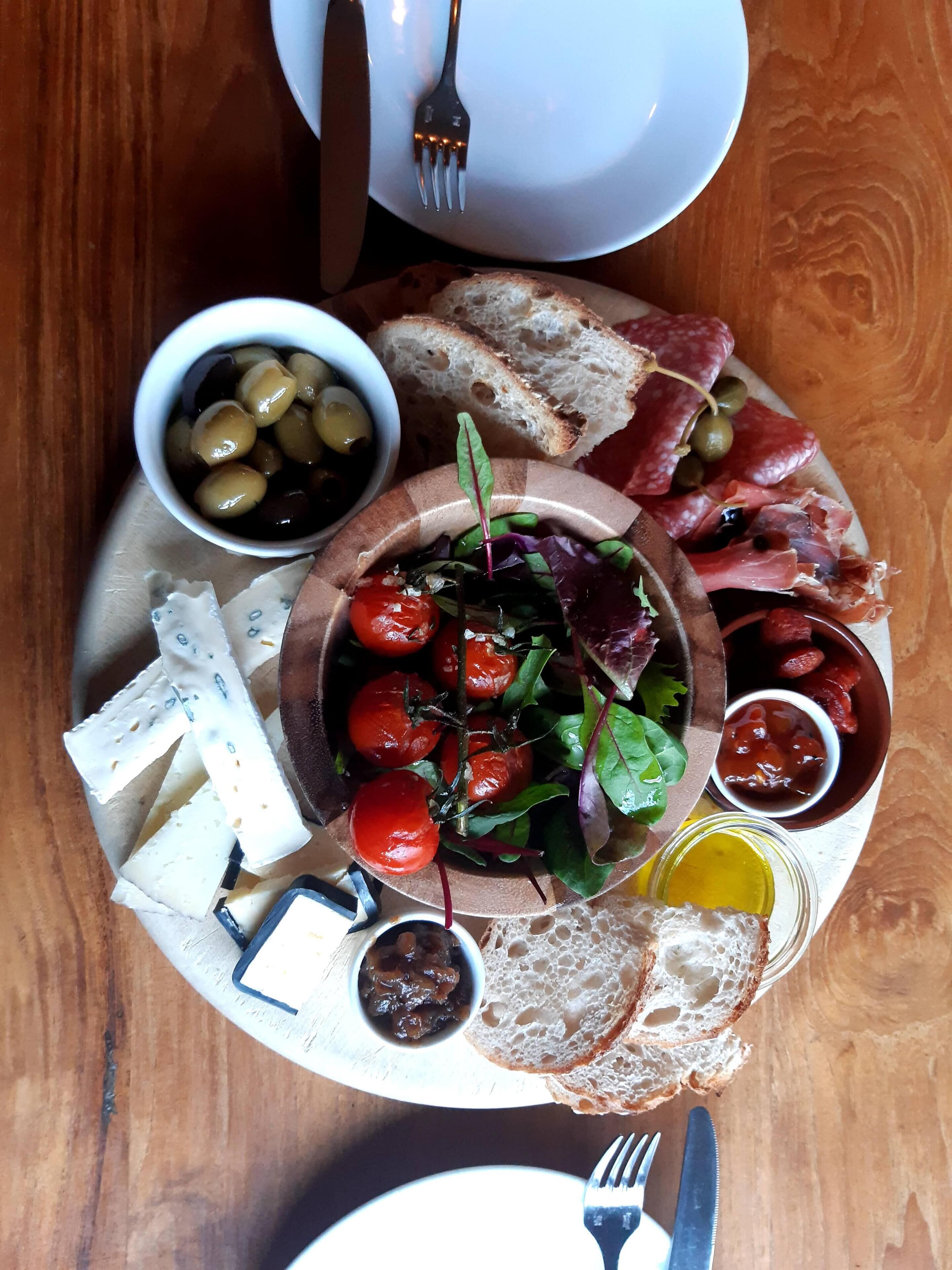 Ein Holztisch mit einer Platte, auf der verschiedene Käsesorten, Oliven, Tomaten und Fleisch angeboten werden.