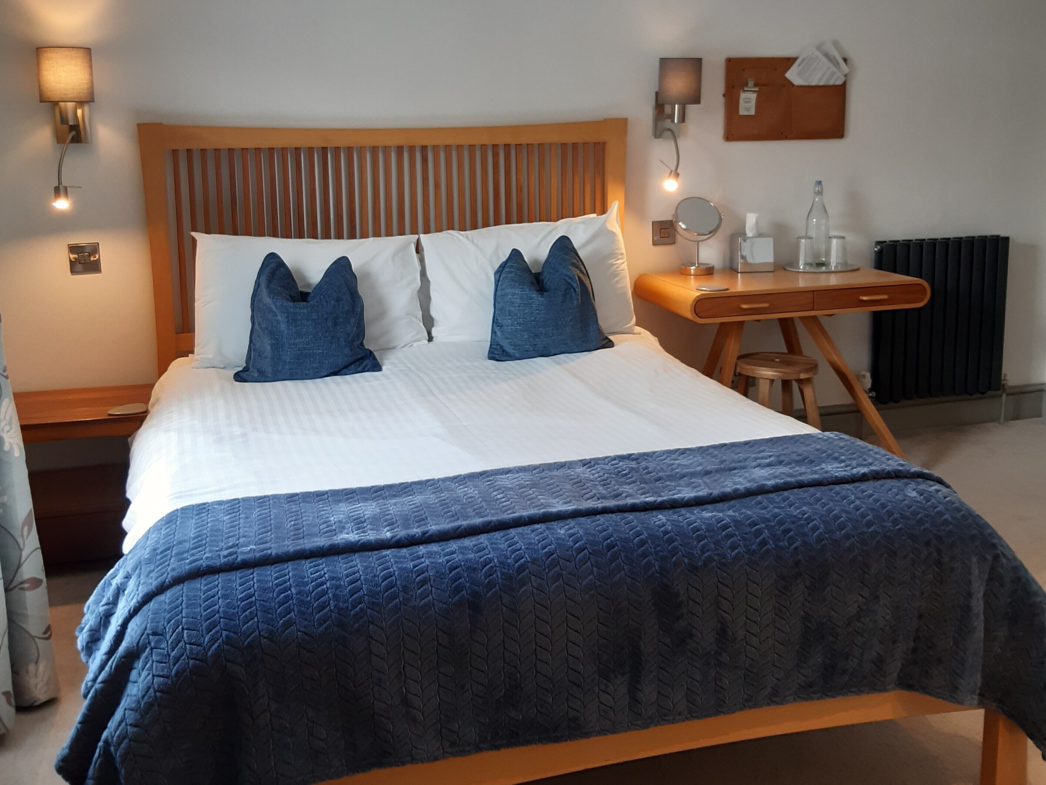Een net opgemaakt bed met een blauw sprei, een houten hoofdbord en twee blauwe kussens. Er staat een nachtkastje met een spiegel, een fles en een pak tissues.