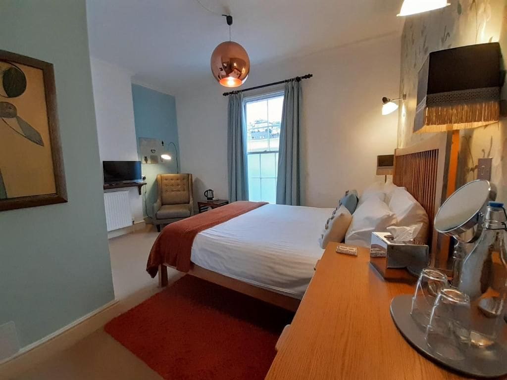 Quarto de hotel com cama, escrivaninha com espelho, cadeira, televisão e janela com cortinas.