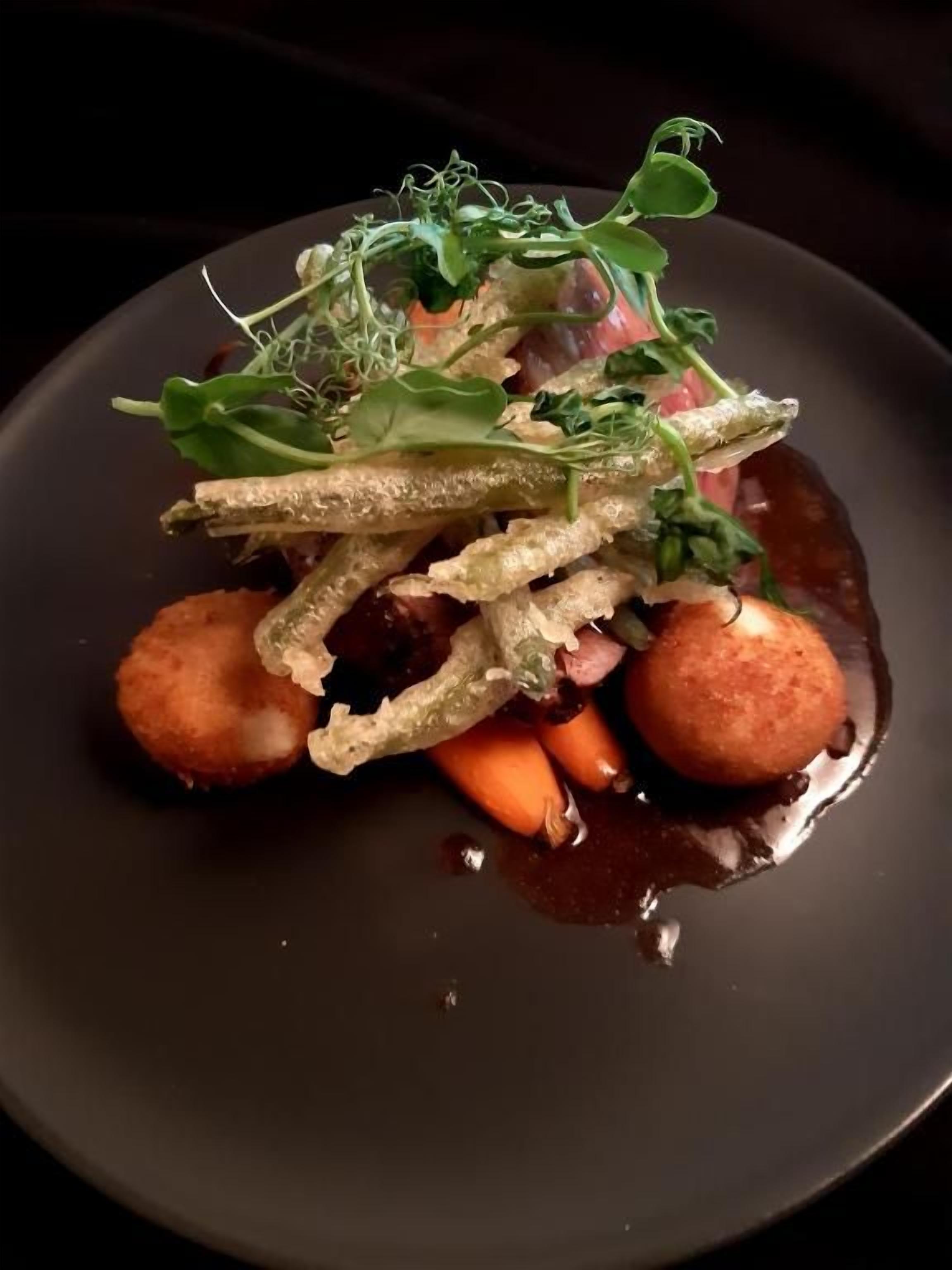 Un plato oscuro con un plato de comida que consiste en esferas, tiras finas y vegetales verdes.