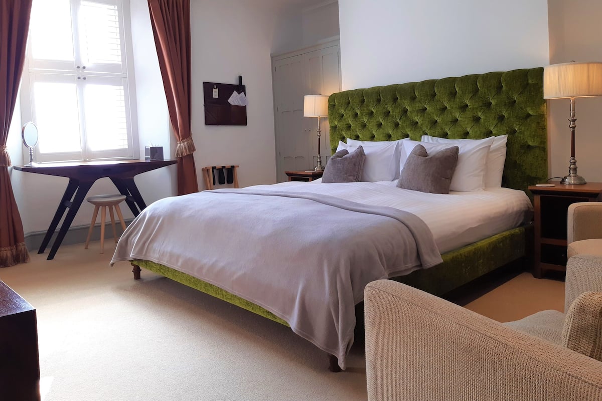 Une chambre d’hôtel avec un grand lit, un bureau, une chaise et une tête de lit recouverte d’un tissu vert.
