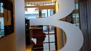 Ein Flur mit einer großen, weißen Skulptur im Stil eines Buchstabens 'B', einem gefliesten Boden und Blick ins Freie durch eine Glastür.
