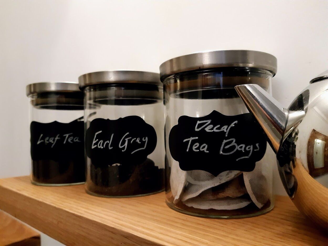 Tre glasbeholdere med metalpropper mærket 'Leaf Tea', 'Earl Grey' og 'Decaf Tea Bags' på en træhylde.