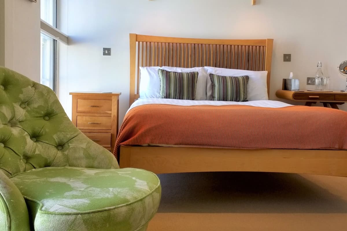 Une chambre à coucher comprenant un lit, un fauteuil à revêtement vert, une table de chevet en bois et deux lampes fixées au mur.