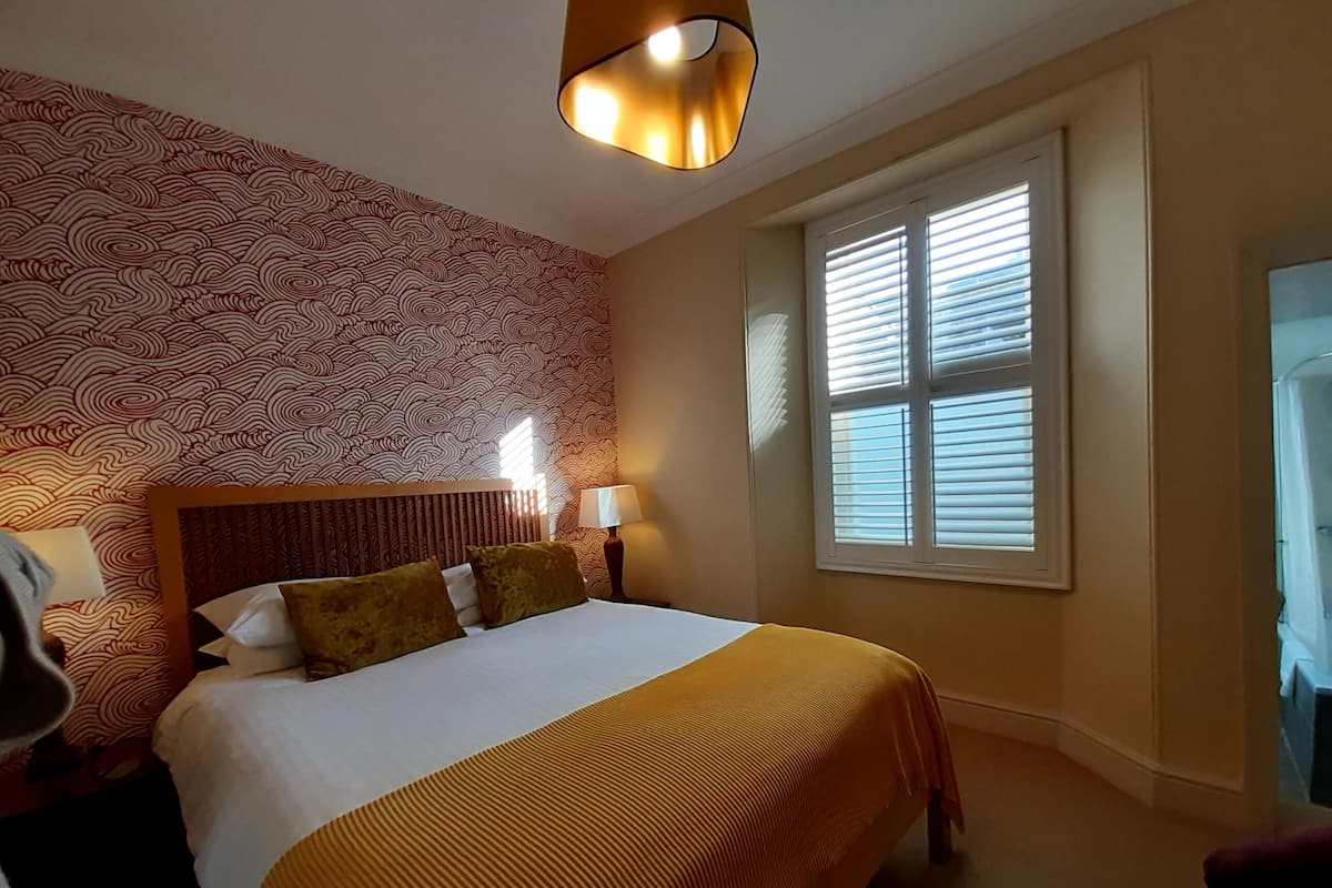 Une chambre d’hôtel avec un grand lit, deux lampes de chevet, un papier peint à motifs et une fenêtre avec des stores.