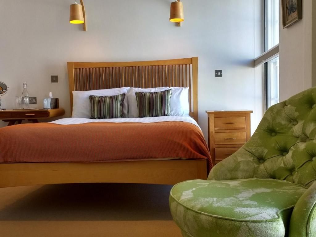 A hotel room with a bed, bedside table, lamp, and a green upholstered chair.