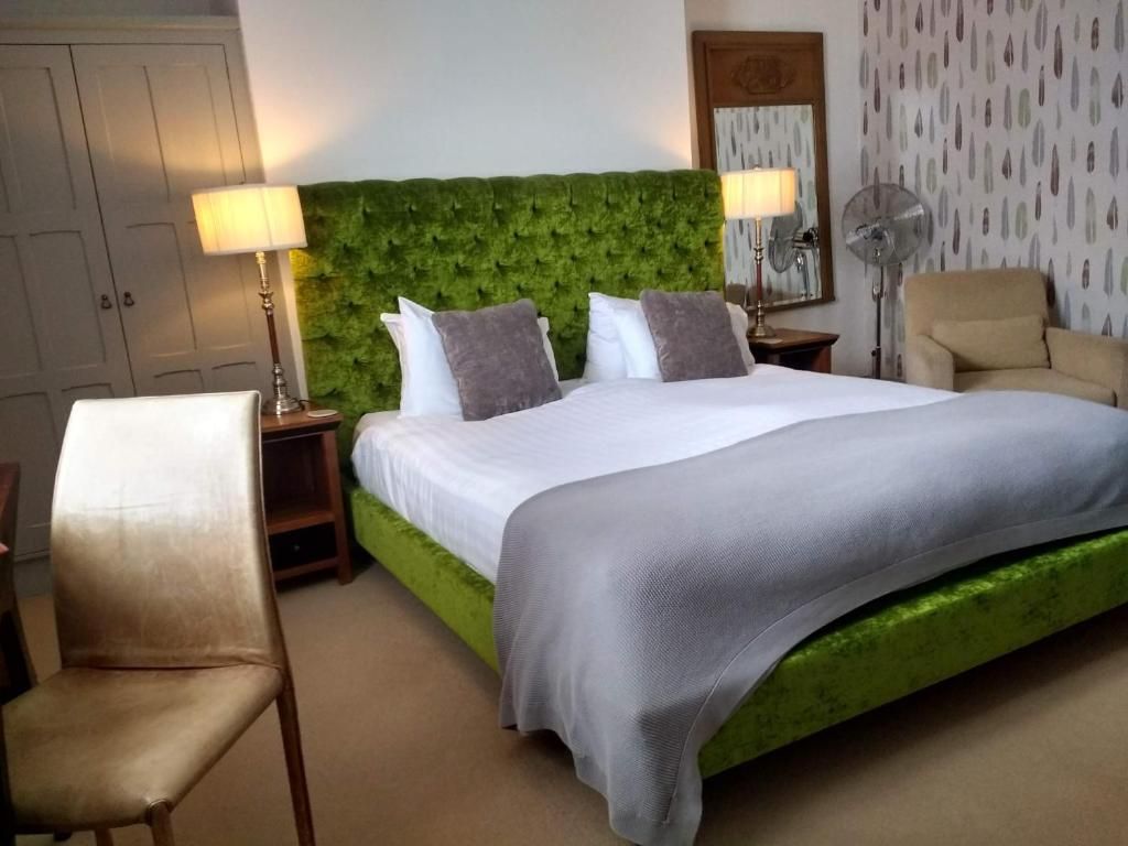 A hotel room with a green upholstered headboard, a large bed with white bedding, two bedside lamps, a chair, and a wall-mounted fan.