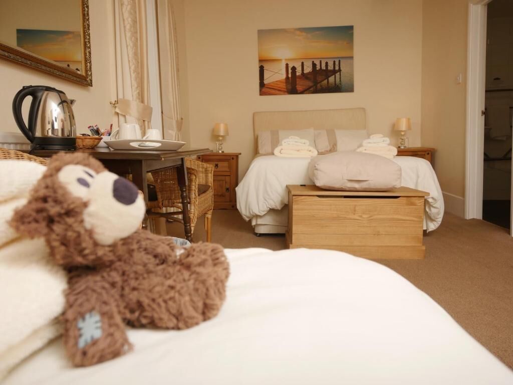 A hotel room with a bed, a desk, a chair, a teddy bear, a painting, and a door.