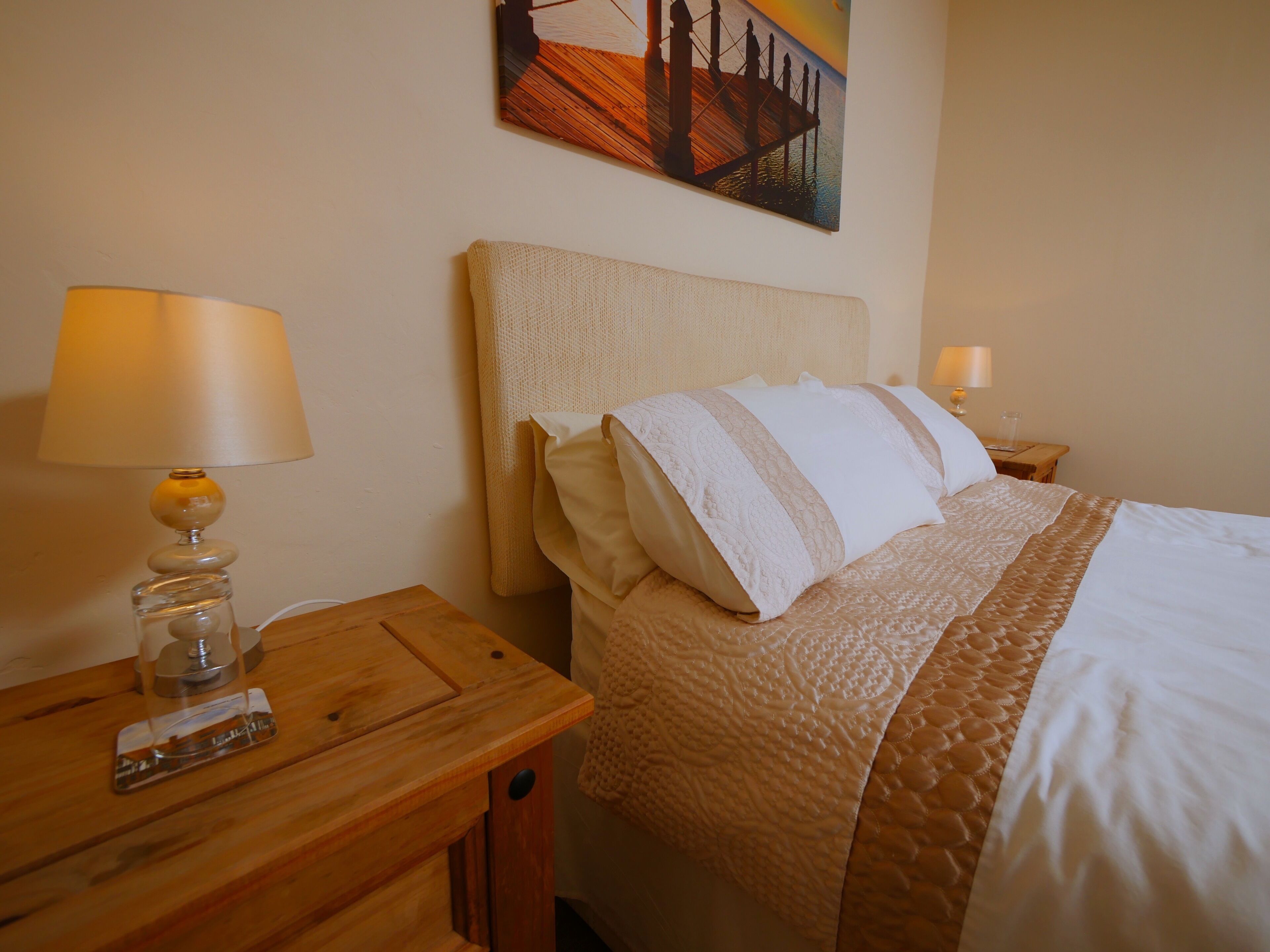 A neatly made bed with a textured blanket and pillows, a wooden nightstand with a lamp, and a painting on the wall.