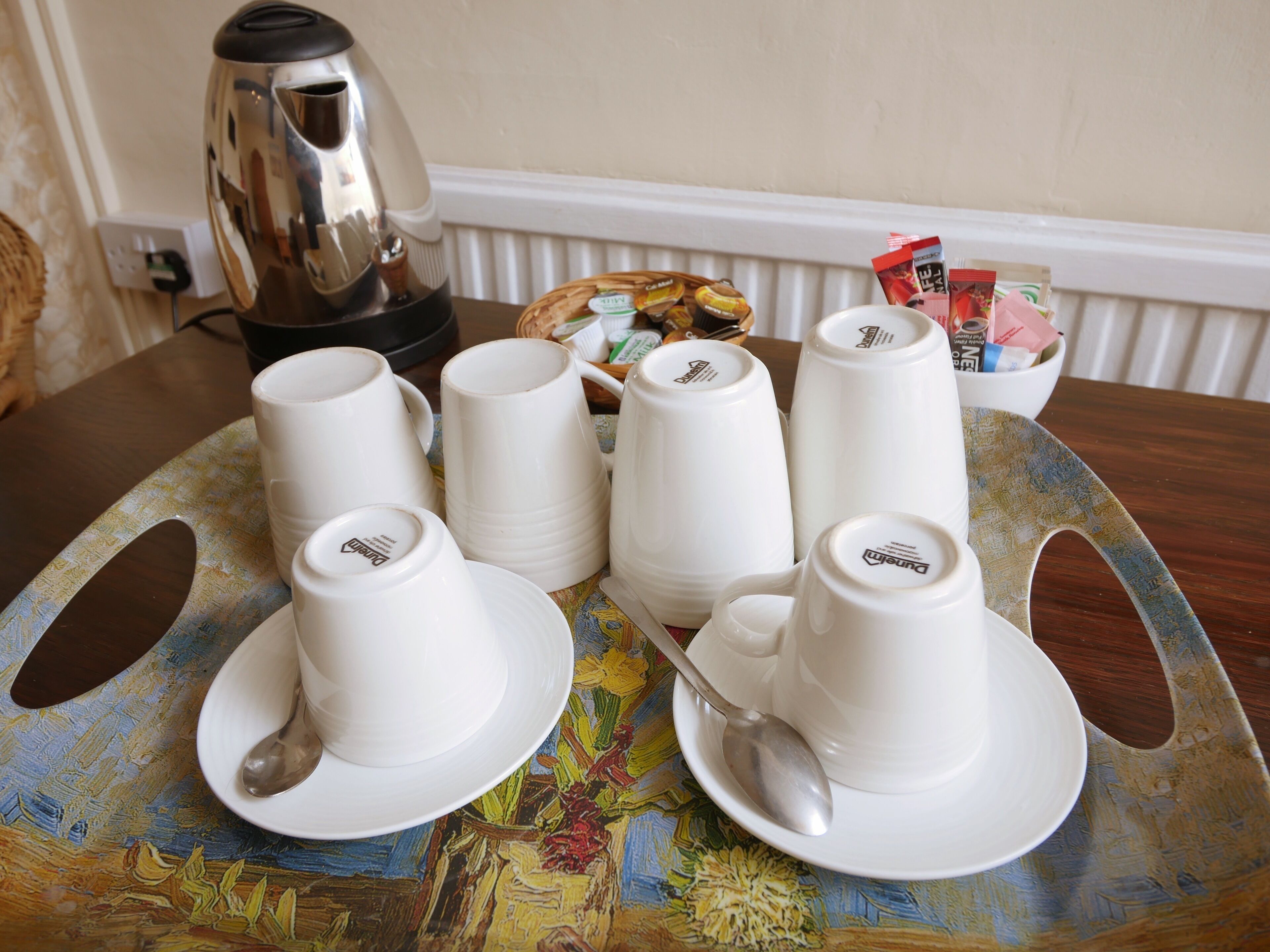 A table with white cups, saucers, and a spoon, a basket of snacks, and a kettle.