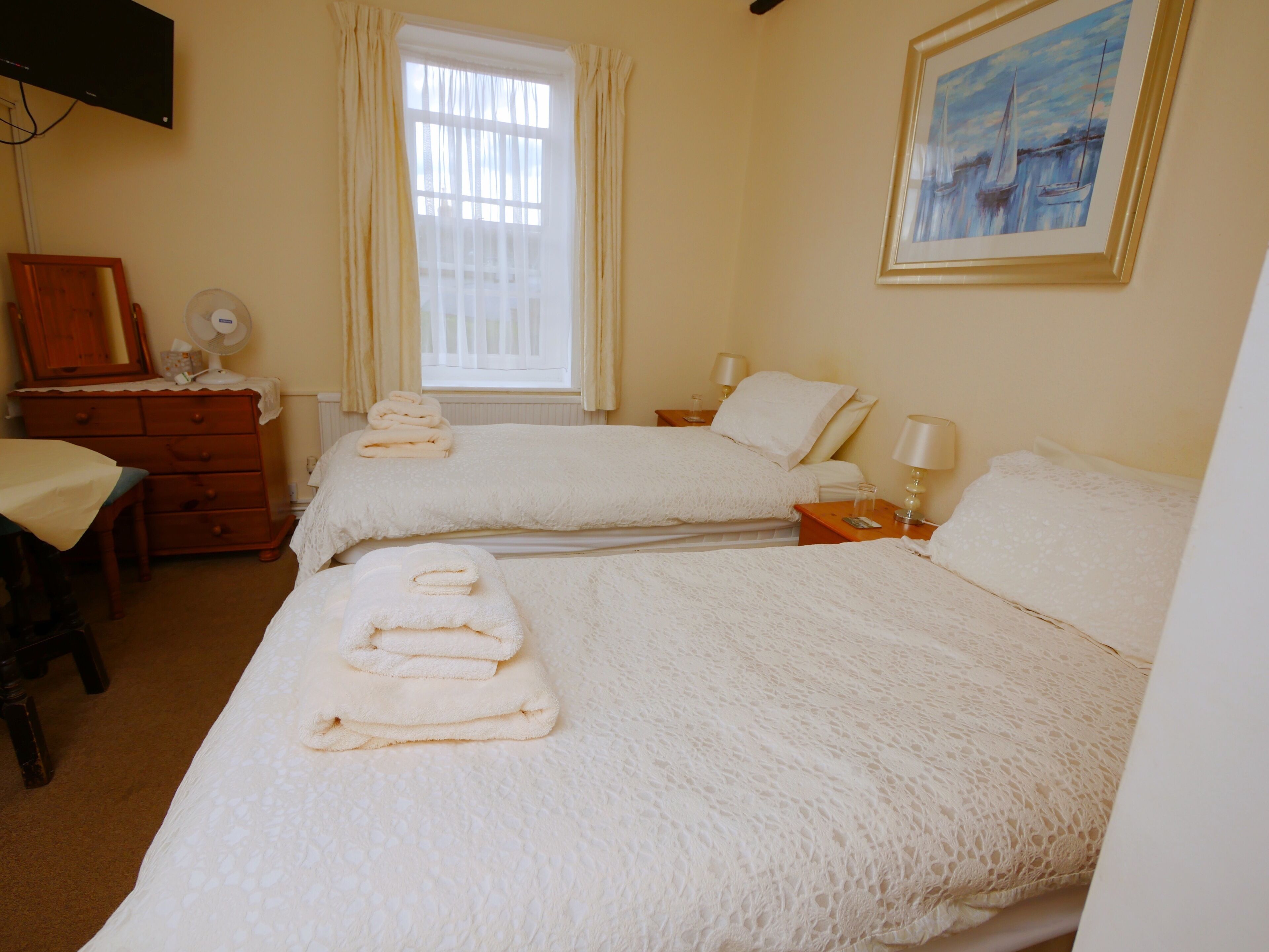 Une chambre d’hôtel avec deux lits, une commode, un bureau, un ventilateur et un tableau au mur.