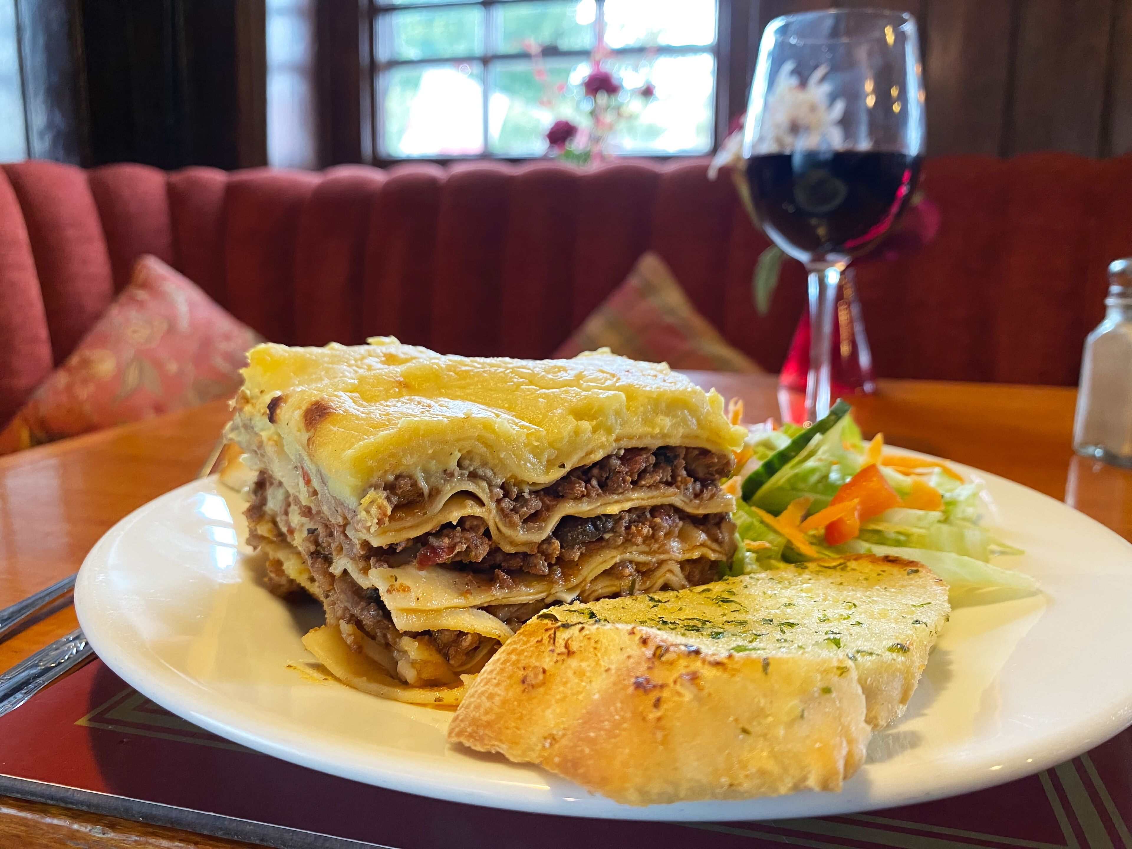 Un plato de guiso de carne y queso en capas, acompañado de una ensalada y una copa de vino tinto sobre una mesa en un restaurante.