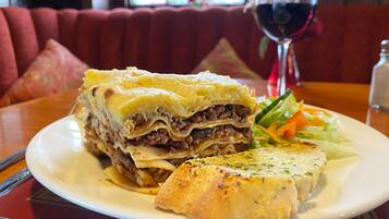 Un plato de guiso de carne y queso en capas, acompañado de una ensalada y una copa de vino tinto sobre una mesa en un restaurante.