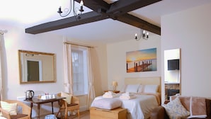 Habitación de hotel con cama, cómoda de madera, silla de mimbre, una mesita con hervidor, sofá y un cuadro de una puesta de sol.