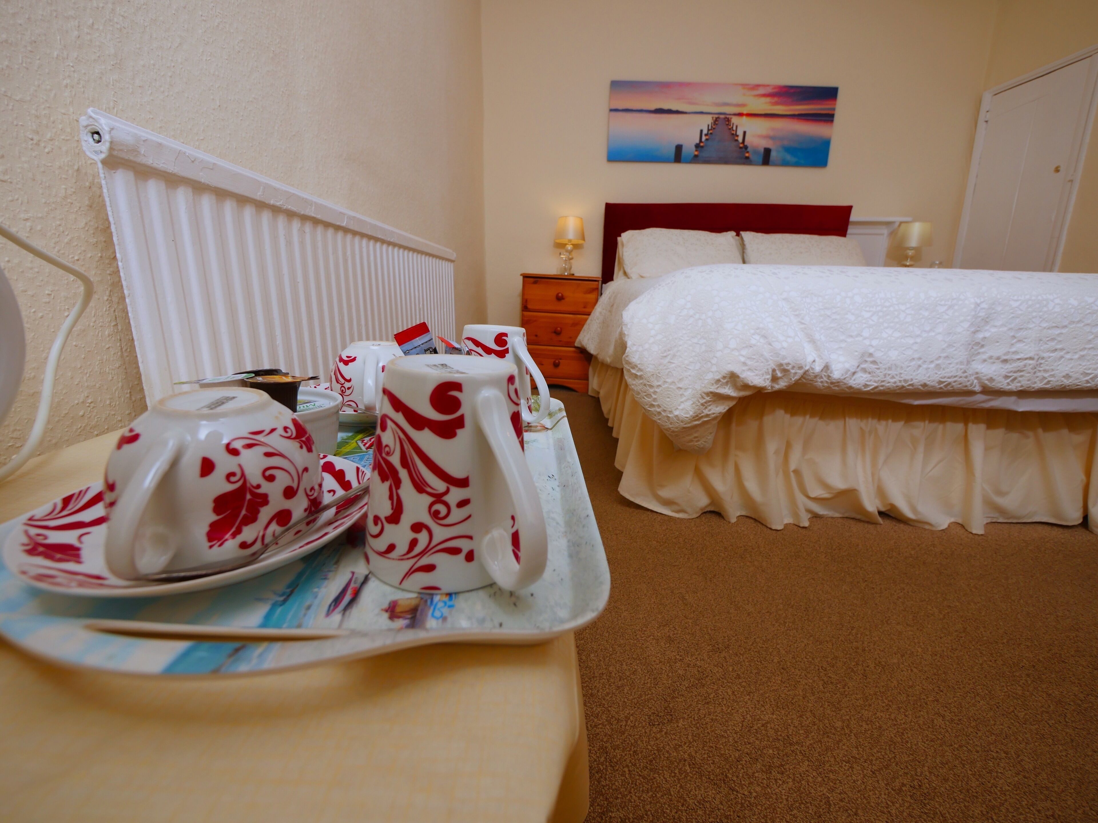 A hotel room with a bed, a bedside table with a tray of breakfast items, a radiator, and a painting on the wall.