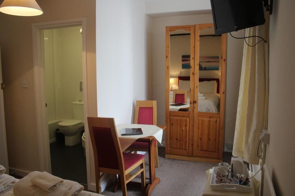 Una habitación con cama, una mesa con sillas, un armario de madera, televisión y un baño visible a través de una puerta abierta.