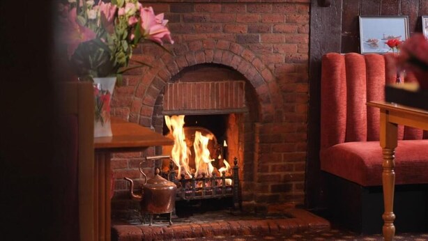 Una chimenea de ladrillo con fuego, una mesa de madera con un florero y un sillón tapizado en rojo.
