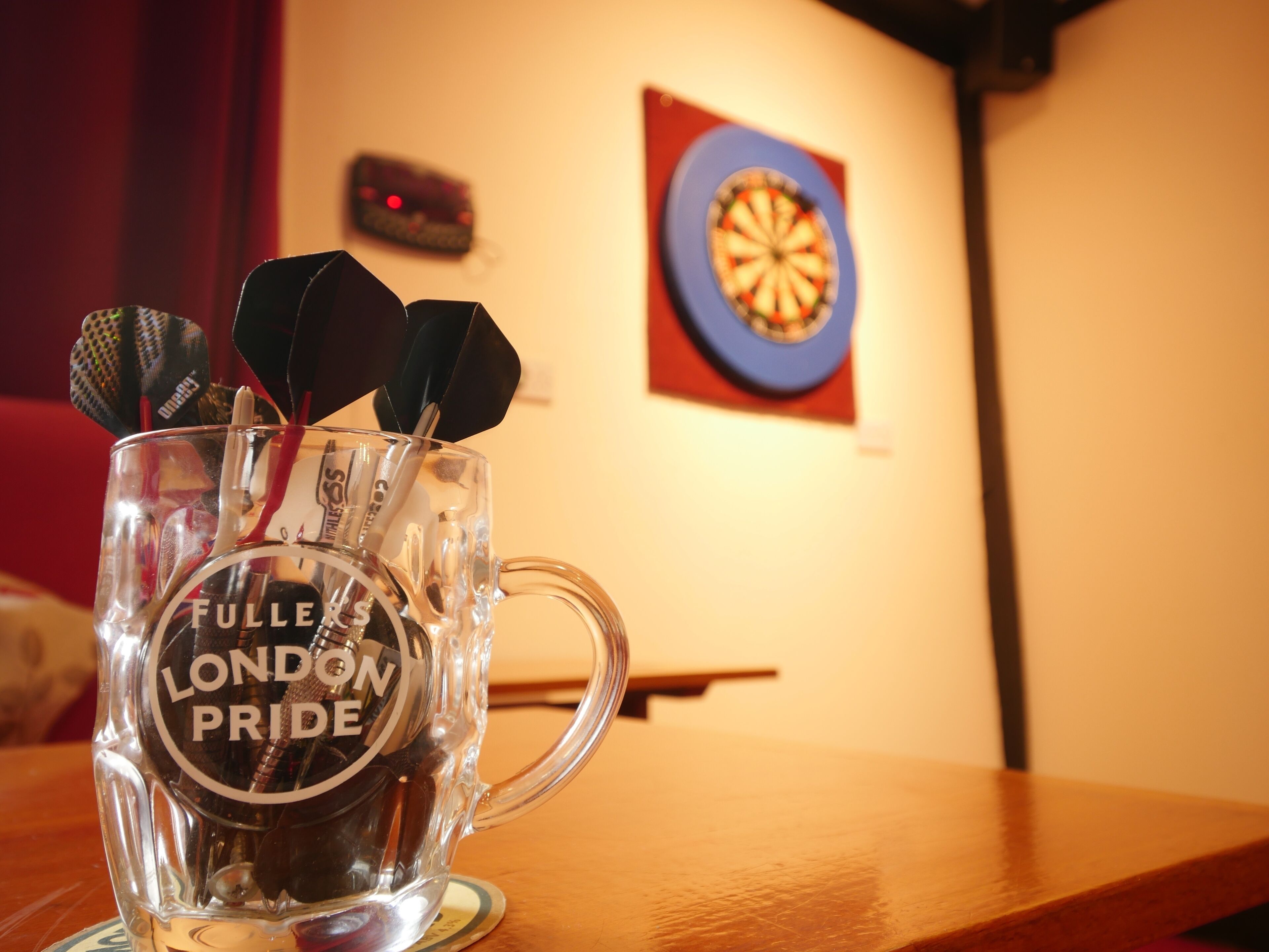 Un vaso de vidrio transparente con el texto "FULLE'S LONDON PRIDE", que contiene dardos y un tablero de dardos al fondo.