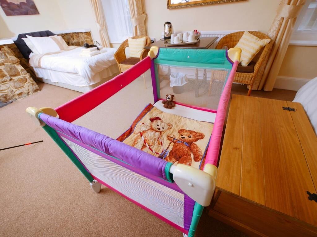 A room with a bed, a small playpen with a teddy bear, and a wicker chair.