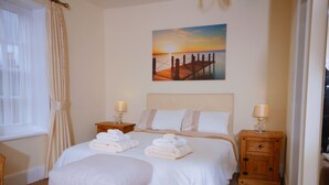 Habitación de hotel con cama, dos mesitas de noche, un banco de madera, un cuadro de un atardecer y una ventana con cortinas.
